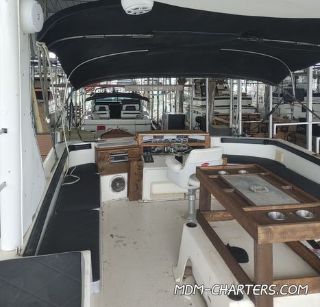 Cozy covered motorboat cockpit in a marina slip with black bimini top, wraparound bench seating, helm station, wooden table with cup holders and other docked boats visible in the background