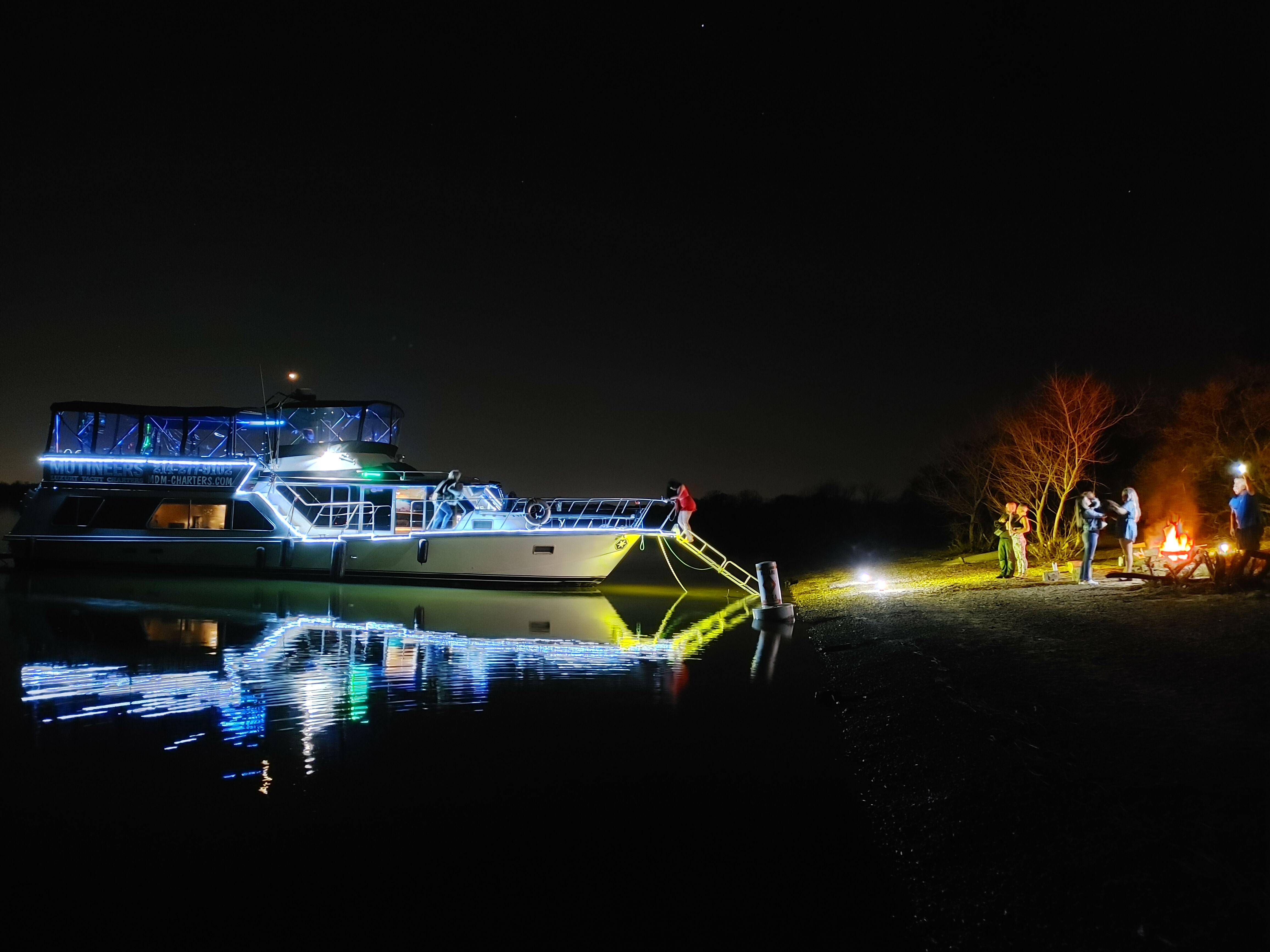 Yacht lit with blue LED accents moored at night, glowing gangway and vivid reflections on calm water, while a small group enjoys a shoreline campfire under a starry sky.