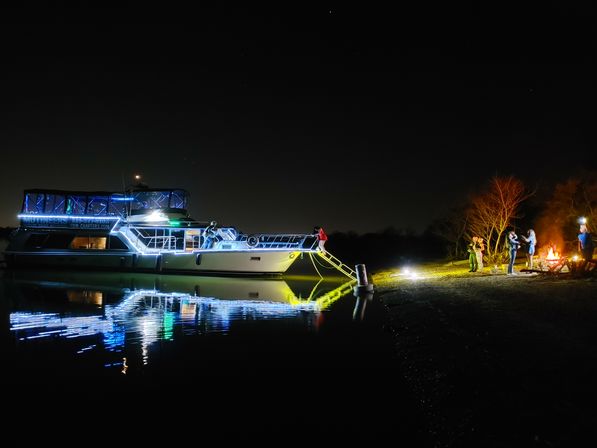 Yacht lit with blue LED accents moored at night, glowing gangway and vivid reflections on calm water, while a small group enjoys a shoreline campfire under a starry sky.