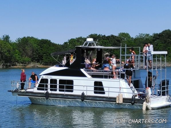 Sunny lake scene showing a multi-level houseboat with people socializing and sunbathing on the upper deck near a tree-lined shore