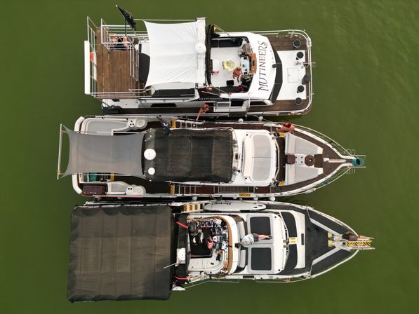 Aerial top-down view of three luxury boats moored side-by-side in green water, showing shaded decks, teak walkways and people relaxing on the yachts