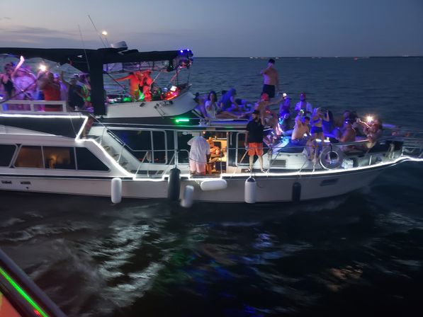 Nighttime yacht party at dusk on open water, groups dancing and lounging under colorful LED and string lights.