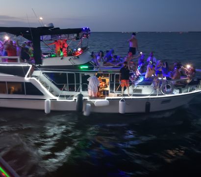 Nighttime yacht party at dusk on open water, groups dancing and lounging under colorful LED and string lights.