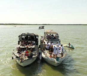 Two motor yachts tied side-by-side on sunny green water with crowds partying on the decks and a nearby jet ski — lively summer boat party on open water