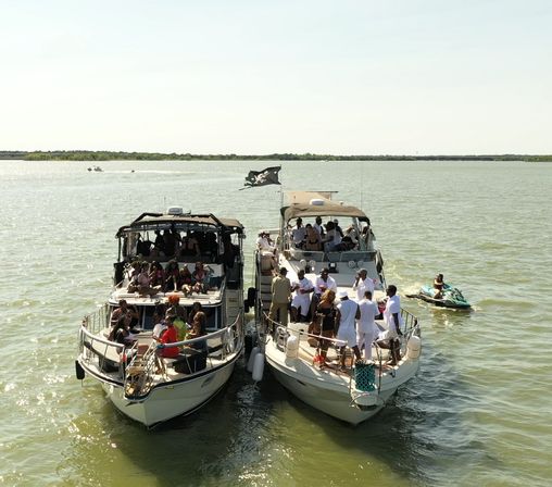 Two motor yachts tied side-by-side on sunny green water with crowds partying on the decks and a nearby jet ski — lively summer boat party on open water