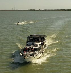 Motor yacht cruising across a sunlit lake with passengers on the foredeck and a jet ski cutting through the wake behind under a clear sky.