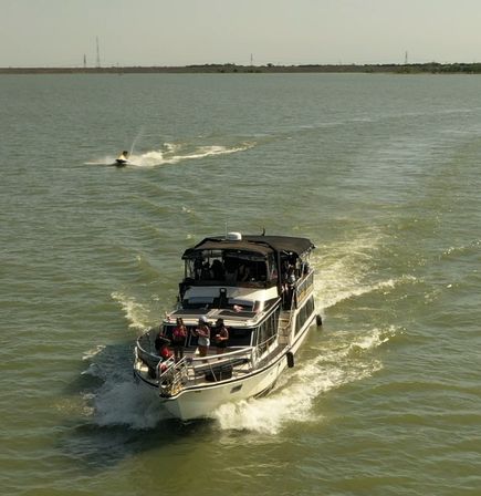 Motor yacht cruising across a sunlit lake with passengers on the foredeck and a jet ski cutting through the wake behind under a clear sky.