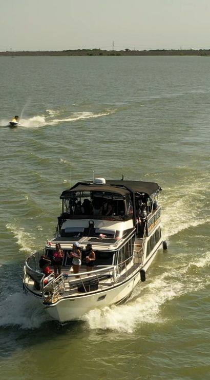 Motor yacht cruising across a sunlit lake with passengers on the foredeck and a jet ski cutting through the wake behind under a clear sky.