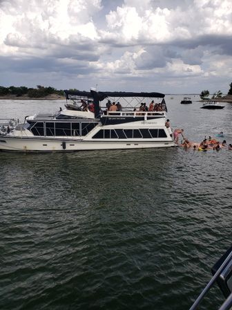 Double-decker party boat anchored on a lake with people swimming and floating near the stern, other small boats nearby under a dramatic cloudy summer sky.