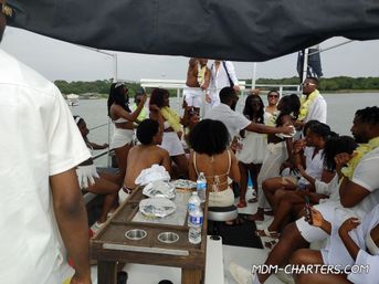Lively summer boat party on a lake — a crowd in white outfits and yellow leis dancing, taking photos and socializing on a party-boat deck with a tree-lined shoreline in the background.