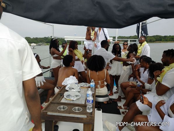 Lively summer boat party on a lake — a crowd in white outfits and yellow leis dancing, taking photos and socializing on a party-boat deck with a tree-lined shoreline in the background.