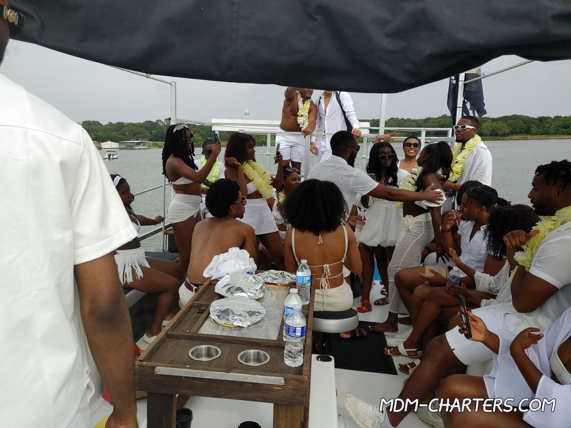 Lively summer boat party on a lake — a crowd in white outfits and yellow leis dancing, taking photos and socializing on a party-boat deck with a tree-lined shoreline in the background.