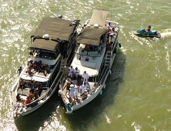 Aerial view of two white motor yachts tied side-by-side hosting a daytime boating party, passengers mingling on the decks and a turquoise jet ski nearby on sunlit green water.