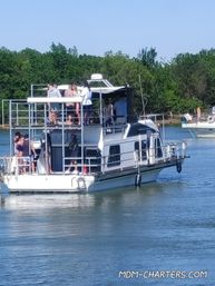Group of people socializing on the upper decks of a two-level houseboat cruising a calm, tree-lined river on a sunny summer day