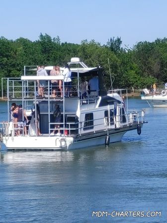 Group of people socializing on the upper decks of a two-level houseboat cruising a calm, tree-lined river on a sunny summer day