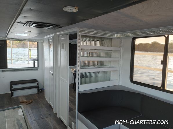 Cozy boat cabin interior with white cabinets, mirrored shelving, black cushioned seating and large windows framing a calm lake shoreline.