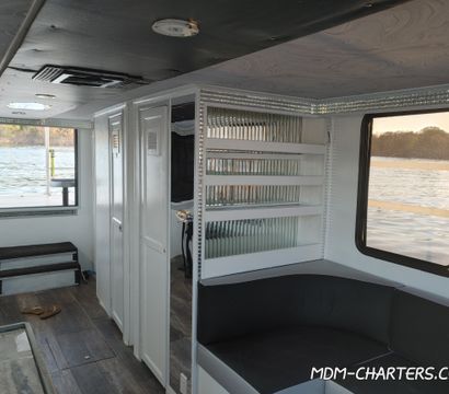 Cozy boat cabin interior with white cabinets, mirrored shelving, black cushioned seating and large windows framing a calm lake shoreline.