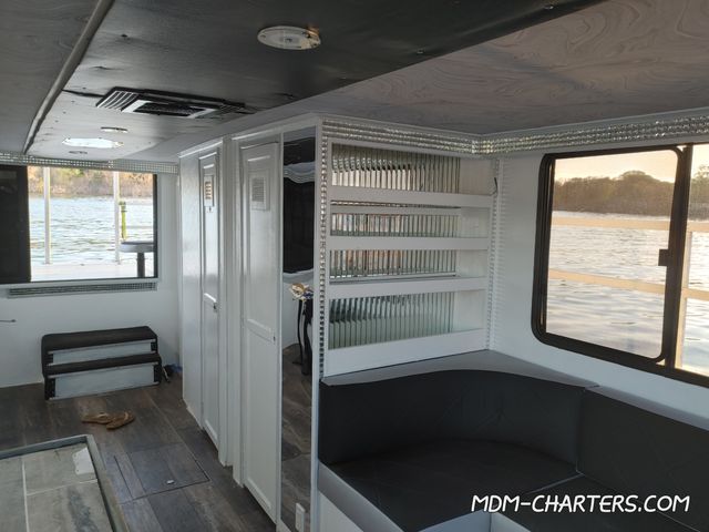 Cozy boat cabin interior with white cabinets, mirrored shelving, black cushioned seating and large windows framing a calm lake shoreline.