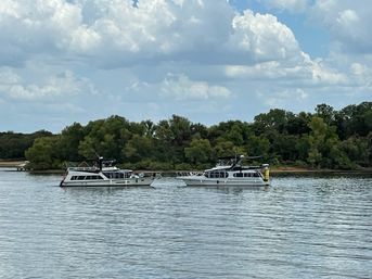 Two white houseboats side-by-side on a calm river near a green tree-lined shore under puffy clouds