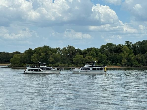 Two white houseboats side-by-side on a calm river near a green tree-lined shore under puffy clouds