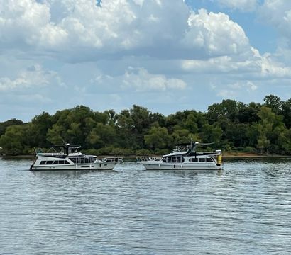 Two white houseboats side-by-side on a calm river near a green tree-lined shore under puffy clouds