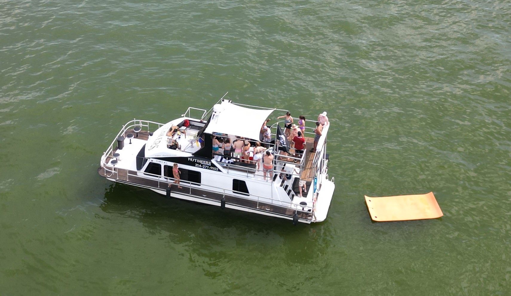 Aerial view of a white party boat on green lake water with people in swimsuits on a shaded upper deck and an orange floating mat nearby.