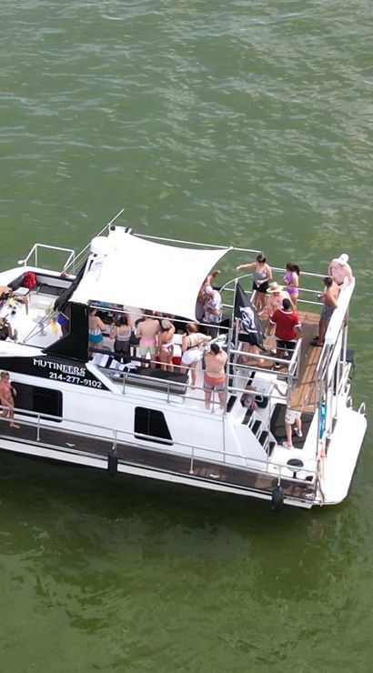 Aerial view of a white party boat on green lake water with people in swimsuits on a shaded upper deck and an orange floating mat nearby.