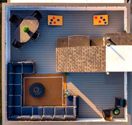 Aerial view of a modern rooftop deck with L-shaped navy sectional around a rug and round fire pit, oval dining table with four chairs and plant centerpiece, two cornhole boards, string lights and extra lounge chairs on blue-gray decking.