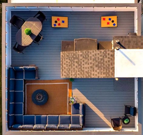 Aerial view of a modern rooftop deck with L-shaped navy sectional around a rug and round fire pit, oval dining table with four chairs and plant centerpiece, two cornhole boards, string lights and extra lounge chairs on blue-gray decking.