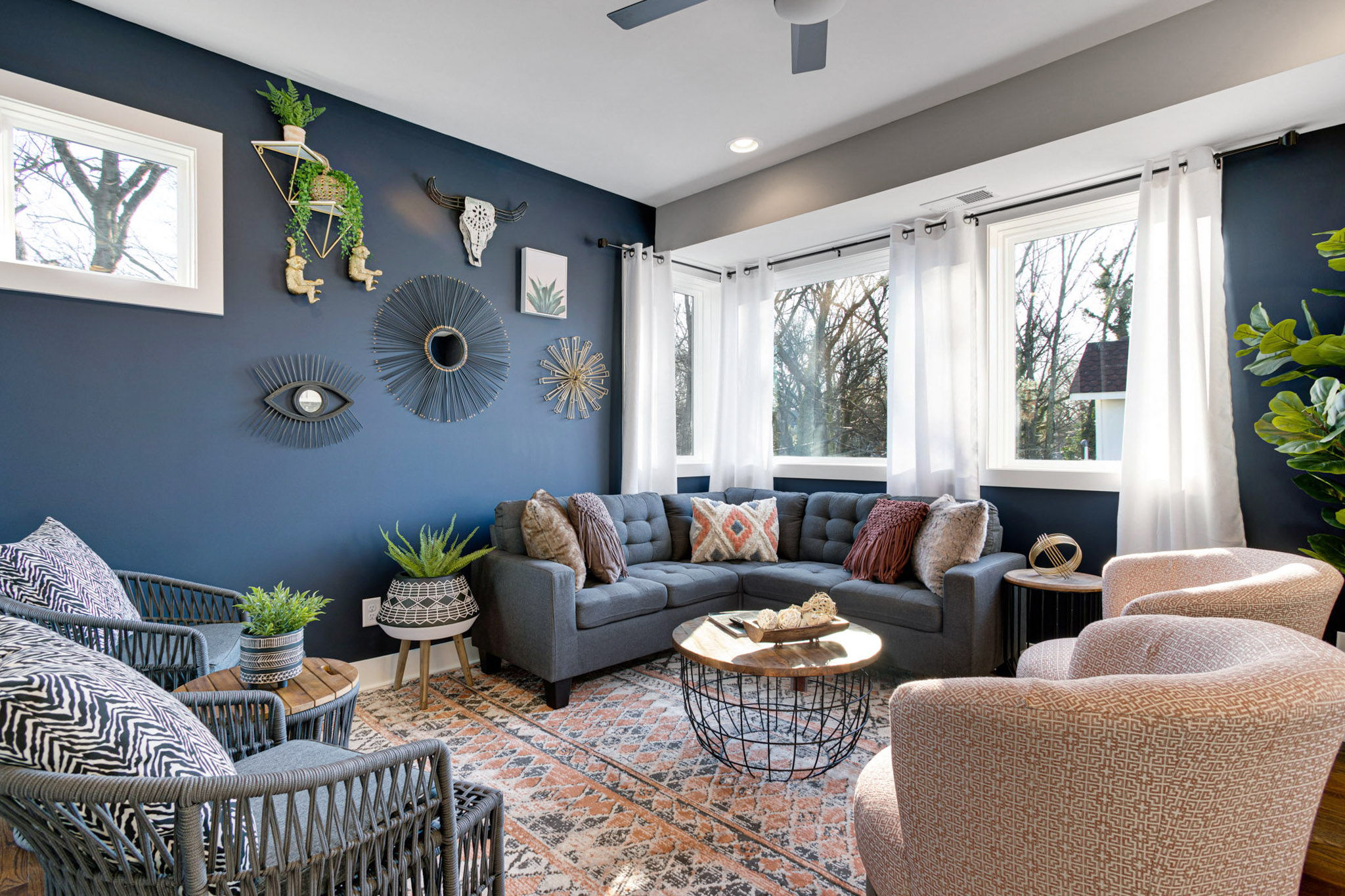 Cozy sunlit living room interior with a navy accent wall, gray sectional sofa, patterned rug, round wire coffee table, mixed seating, decorative wall art and indoor plants.