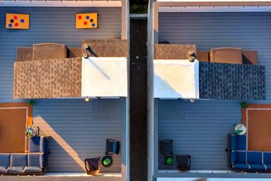 Aerial view of twin sunlit rooftop decks on modern townhouses with gray decking, blue patio sofas, small tables, planters and a mirrored layout.