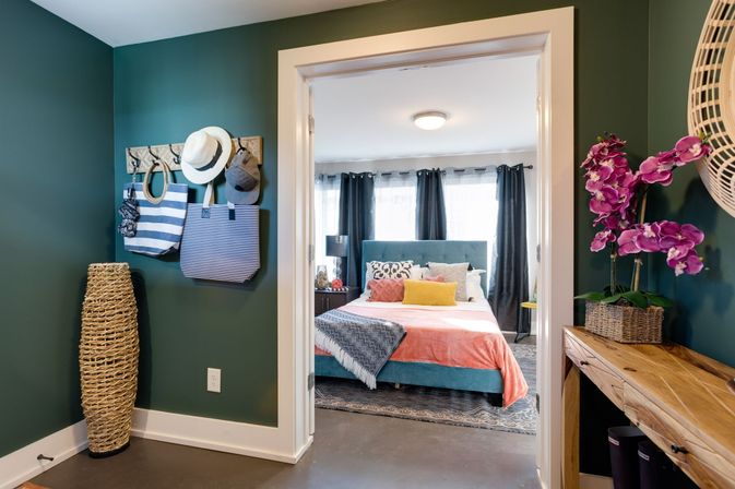 Home entryway with dark teal walls, striped tote bags and hats on hooks, tall woven floor vase and a wooden console with magenta orchids, opening into a bright bedroom with a teal upholstered bed, coral throw, patterned pillows and dark curtains.