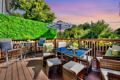 Cozy suburban backyard deck at sunset with wicker dining set, cushioned chairs and stools, striped patio umbrella, colorful throw pillows, potted plants, green privacy hedge and string lights for al fresco dining.