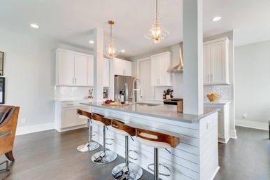 Bright open-concept kitchen with white shaker cabinets, long gray island topped with three wood-and-chrome bar stools, clear glass pendant lights, stainless steel appliances, and herringbone tile backsplash.