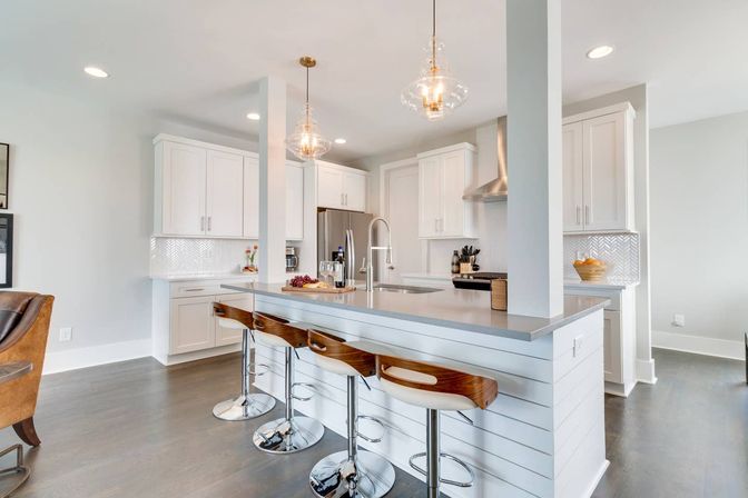 Bright open-concept kitchen with white shaker cabinets, long gray island topped with three wood-and-chrome bar stools, clear glass pendant lights, stainless steel appliances, and herringbone tile backsplash.