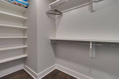 Modern residential walk-in closet with white built-in shelves, chrome clothing rods, a few metal hangers, gray walls and dark hardwood floor.