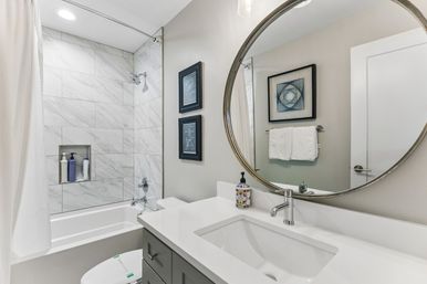 Sleek modern bathroom with white marble-tile shower and tub, built-in shampoo niche, gray vanity, round mirror and chrome fixtures.