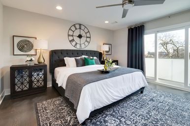 Modern master bedroom with king bed and tufted dark headboard, oversized wall clock, gray-and-white bedding, patterned rug, nightstands with lamps, and floor-to-ceiling windows opening to a balcony with tree-lined view.