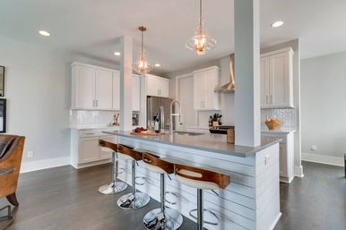 Bright modern open-concept kitchen — white cabinets, gray island with curved wood bar stools, glass pendant lights, stainless appliances and herringbone tile backsplash