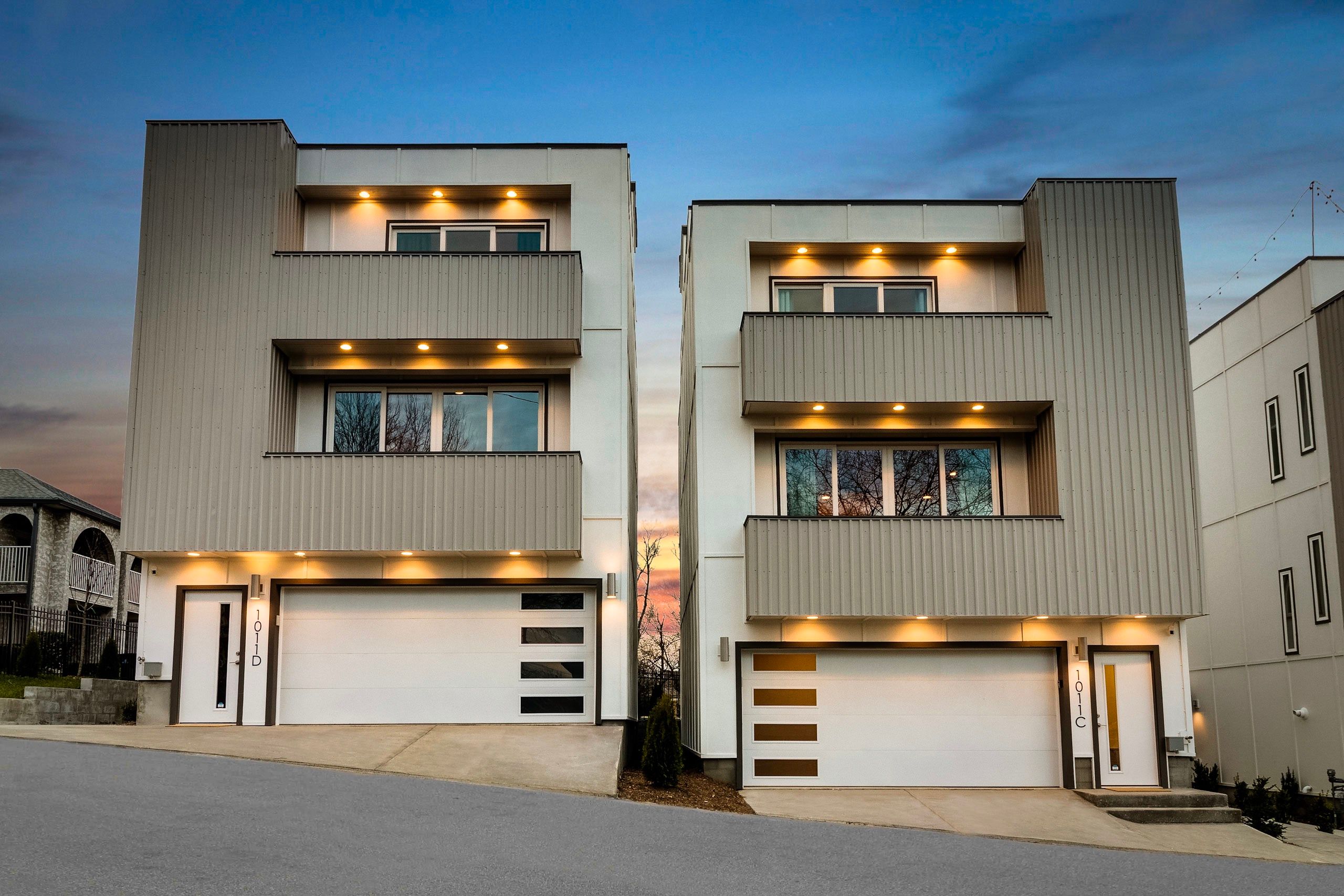 Twin modern three-story townhouses with balconies, attached white garage doors and warm recessed exterior lights under a sunset sky on a sloped urban residential street