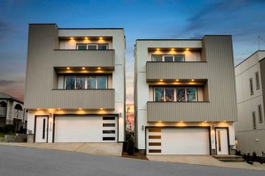 Twin modern three-story townhouses with balconies, attached white garage doors and warm recessed exterior lights under a sunset sky on a sloped urban residential street