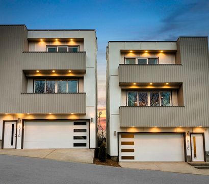Twin modern three-story townhouses with balconies, attached white garage doors and warm recessed exterior lights under a sunset sky on a sloped urban residential street