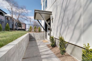 Basement-level side entrance of a modern townhouse with a concrete retaining wall, narrow walkway to a covered door with angled awning, small shrubs and iron fence under a clear blue sky.