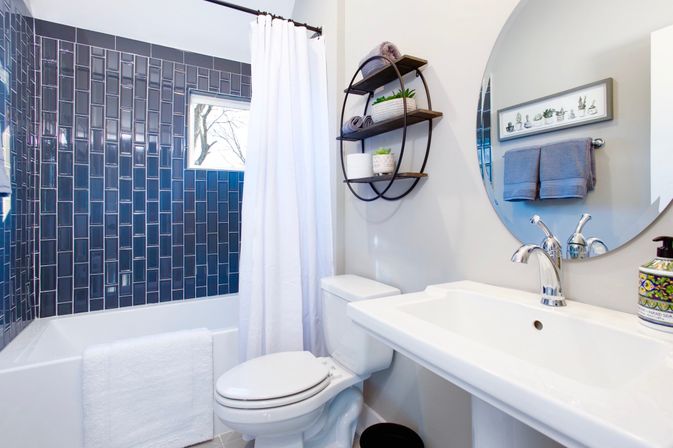 Modern residential bathroom with glossy navy subway-tile shower wall, white tub and curtain, pedestal sink with chrome faucet, round mirror, circular wood-and-metal wall shelves with potted plants, and white toilet.