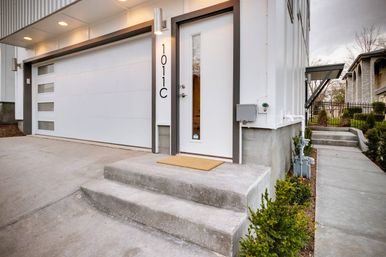 Modern white townhouse entrance with sleek garage door featuring frosted windows, concrete steps, tan doormat, and neat shrubs along a suburban sidewalk.