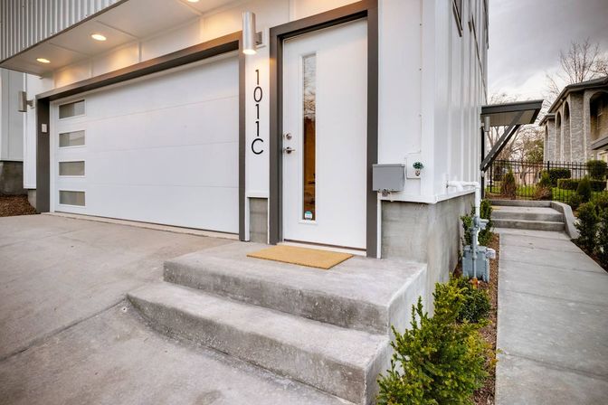 Modern white townhouse entrance with sleek garage door featuring frosted windows, concrete steps, tan doormat, and neat shrubs along a suburban sidewalk.