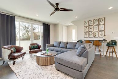 Bright modern living room with large sliding glass doors and dark curtains, roomy gray sectional, two brown leather accent chairs, round wooden coffee table on patterned rug, bar stools, botanical wall art and ceiling fan