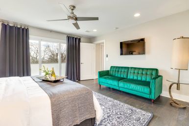 Modern bedroom interior with king bed and gray throw, floor-to-ceiling windows with dark curtains opening to a balcony view of trees, emerald green velvet sofa, wall-mounted TV, ceiling fan, patterned rug and a tray with wine on the bed.