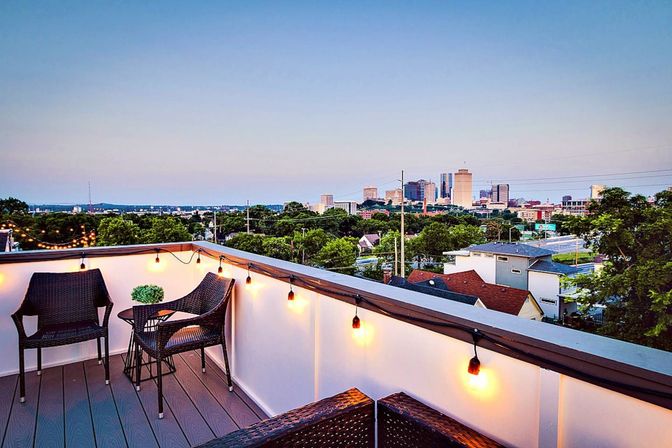 Rooftop patio with wicker chairs, small table and warm string lights overlooking a tree-lined neighborhood and distant downtown skyline at dusk