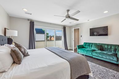 Bright modern bedroom with a king-size bed in white and gray linens, emerald velvet sofa, wall-mounted TV, ceiling fan and sliding glass doors leading to a small balcony with rooftop views.